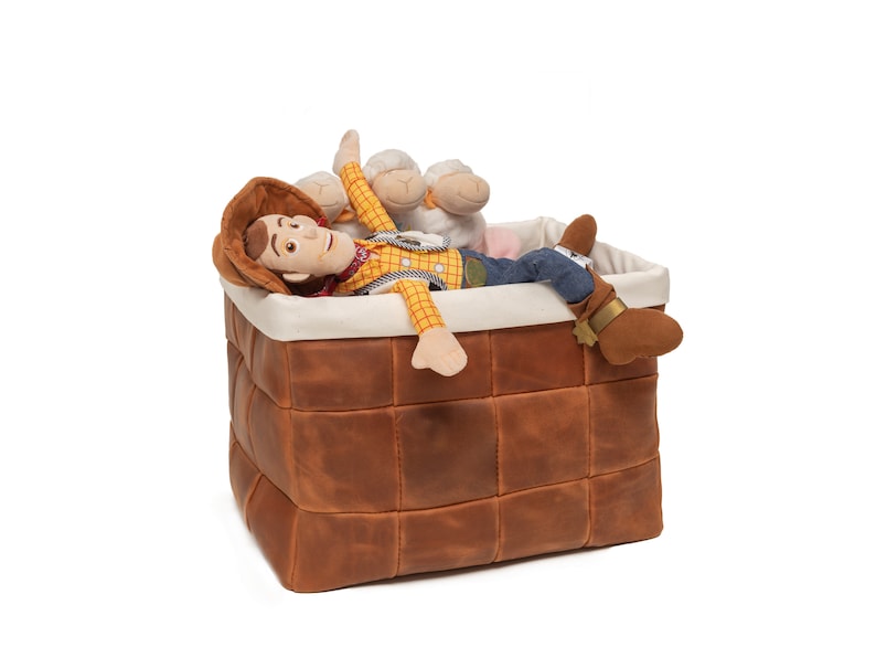 May include: A brown leather storage basket with a white lining. A toy cowboy doll with a yellow shirt and blue jeans is lying inside the basket. Two white plush sheep toys are also in the basket.