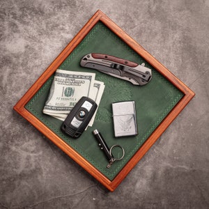 Könnte beinhalten: Ein grünes Ledertablett mit einem Holzrahmen, das einen Stapel US-Dollar-Scheine, einen Autoschlüssel, ein silbernes Feuerzeug mit Adlermotiv und ein Klappmesser mit Holzgriff enthält.