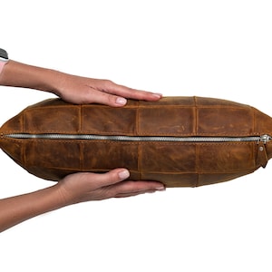 May include: A brown leather pillow with a zipper closure. The pillow is made of multiple pieces of leather sewn together.