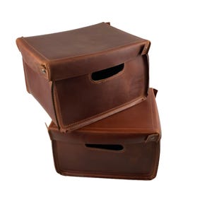 May include: Two stacked, brown leather storage boxes with cut-out handles. The boxes have a rectangular shape with a hinged lid and are secured with metal rivets. The leather has a slightly distressed appearance.