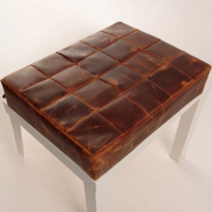 May include: A rectangular ottoman featuring a top surface constructed from a grid of stitched brown leather squares with a distressed finish. The seat is supported by four straight, solid white legs positioned at each corner of the base.