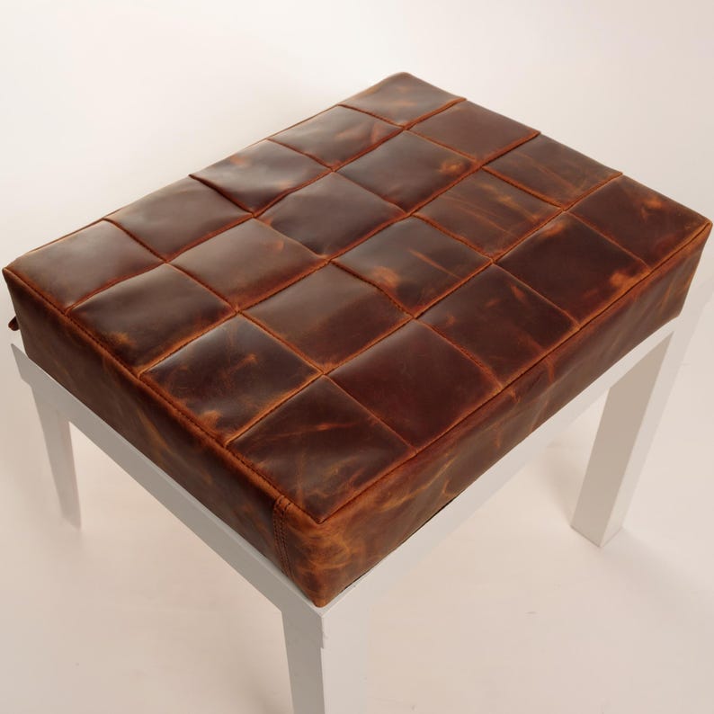 May include: A rectangular ottoman featuring a top surface constructed from a grid of stitched brown leather squares with a distressed finish. The seat is supported by four straight, solid white legs positioned at each corner of the base.