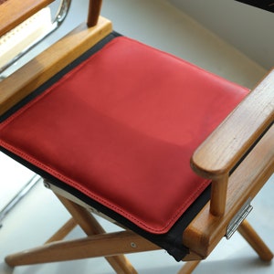 May include: A wooden director's chair with a red leather seat cushion. The chair has a black canvas back and wooden arms.