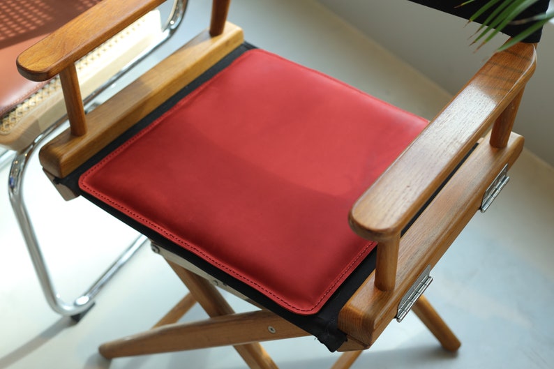 May include: A wooden director's chair with a red leather seat cushion. The chair has a black canvas back and wooden arms.
