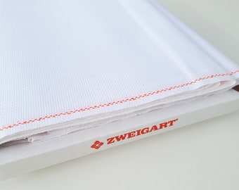 Puede incluir: Tela Aida blanca doblada sobre una caja blanca con el texto rojo "ZWEIGART" impreso en la caja.