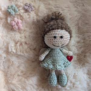 Könnte beinhalten: Eine gehäkelte Puppe mit braunem Garnhaar in einem Dutt, bekleidet mit einem hellgrünen Kleid mit gerüschtem Saum und passenden Schuhen. Die Puppe hat schwarze Perlenaugen, ein gesticktes Lächeln und hält ein kleines rotes Herz. Drei winzige gehäkelte Blumen in Minze, Rosa und Lavendel liegen daneben.