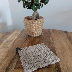 Könnte beinhalten: Ein quadratischer, beigefarbener Makramee-Topflappen mit einer dunkelbraunen Lederschlaufe liegt auf einem Holztisch. Ein kleiner Bonsai in einem geflochtenen Korb steht im Hintergrund. Das Makramee hat ein sich wiederholendes Sternmuster.