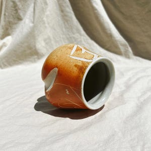 May include: A handmade ceramic mug with a warm, orange-brown glaze. The mug features white, geometric designs and a light gray interior rim. The mug is resting on a neutral-colored, textured surface.