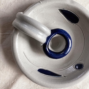 May include: A white ceramic candle holder with a blue ring and blue leaf designs. The holder has a handle on the side.