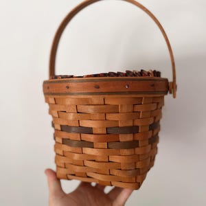 May include: A woven basket with a curved handle. The basket is made of light brown wood with darker brown accents. The handle is attached to the top rim of the basket. The basket is held by a hand.