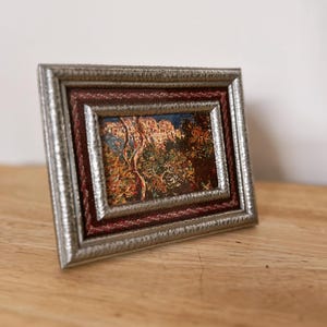 May include: A small framed painting with a silver-colored textured frame and a burgundy rope-like inner border. The painting depicts a landscape with trees and buildings, featuring warm colors.