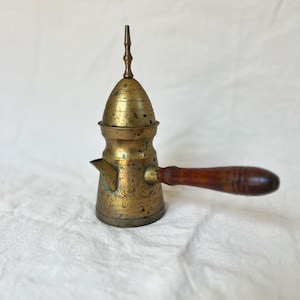May include: A vintage brass coffee pot with a wooden handle. The pot has an ornate design etched into the metal and a long, slender spout. The handle is a dark brown color. The pot is approximately 6 inches tall.