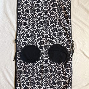 May include: A black and white patterned apron with a floral design. The apron has two large, round black pockets and black trim. The apron is tied with black strings.