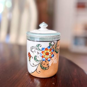 May include: A small, lidded ceramic jar with a floral design. The jar is white with a light orange base and a blue and white lid. The floral design features a large flower with blue, orange, and yellow petals, and green leaves.