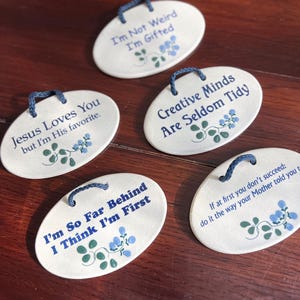 May include: Five oval ceramic plaques with blue rope hangers. Each plaque has a different inspirational quote in blue text, with floral and leaf accents. The quotes include: "I'm Not Weird I'm Gifted" and "Jesus Loves You but I'm His favorite."