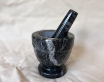 May include: A small, dark gray mortar and pestle set. The mortar is a bowl shape with a rounded base, and the pestle is cylindrical. The set is made of stone with a marbled pattern. The background is a light-colored, textured fabric.