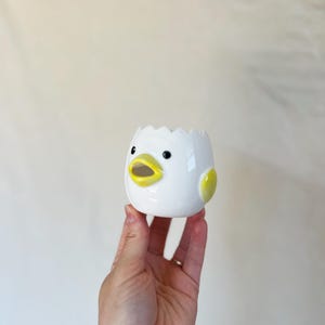 May include: A white ceramic eggshell-shaped planter with a chick face design. The planter has black eyes, a yellow beak, and yellow accents on the sides. The top edge of the eggshell is jagged. The planter is held in a hand.