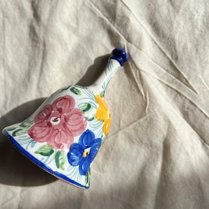 May include: A white ceramic bell with a blue rim and floral design. The bell is painted with pink, yellow, blue, and green flowers.
