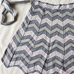 May include: A vintage-style apron with a chevron pattern in gray, white, and pink. The apron features a scalloped hem and long ties for securing around the waist. The fabric has a floral and geometric design.