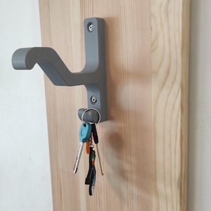 Könnte beinhalten: Ein grauer Schlüsselhalter an einer hellen Holzplatte befestigt. Der Schlüsselhalter hat ein gebogenes Design und hält einen Satz Schlüssel mit verschiedenen Schlüsselanhängern. Die Holzmaserung ist sichtbar, und der Schlüsselhalter ist mit Schrauben befestigt.