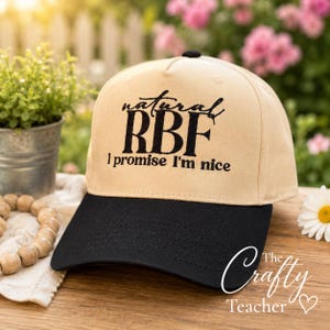 May include: A beige baseball cap with a black peak. The cap has the embroidered text "natural RBF, I promise I'm nice" in black. A small black button is on top.