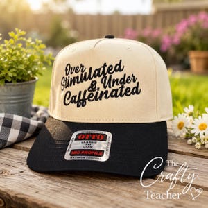 Puede incluir: Gorra de béisbol beige y negra con el texto bordado "Over Stimulated & Under Caffeinated". La gorra tiene una visera negra y un botón negro en la parte superior. La gorra está sobre una superficie de madera.