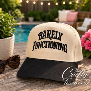 May include: A beige and black baseball cap with the text "BARELY FUNCTIONING" embroidered in black. The cap is sitting on a wooden surface next to sunglasses and a potted plant. The Crafty Teacher logo is on the side.