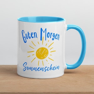 May include: White ceramic mug with a blue rim and handle. The mug has a yellow sun with blue rays and the text "Guten Morgen Sonnenschein" in blue.