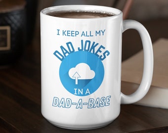 May include: White ceramic mug with the text "I KEEP ALL MY DAD JOKES IN A DAD-A-BASE" in blue. The design includes a cloud icon with an upward arrow. The mug is on a dark wooden surface.