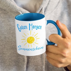 May include: A white mug with a blue rim and handle. The mug has a yellow sun and the text "Guten Morgen Sonnenschein" in blue.