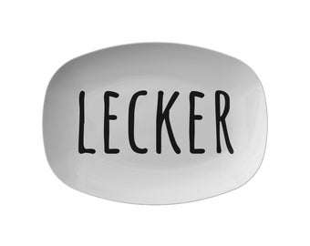 May include: A white, oval-shaped serving platter with the word "LECKER" printed in bold, black letters. The platter has a glossy finish and a simple, modern design, suitable for serving food.