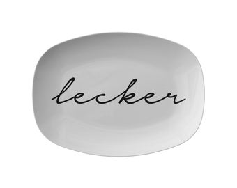 May include: A white, rectangular plate with rounded corners. The word "lecker" is written in black cursive across the center of the plate. The plate has a glossy finish and is set against a white background.