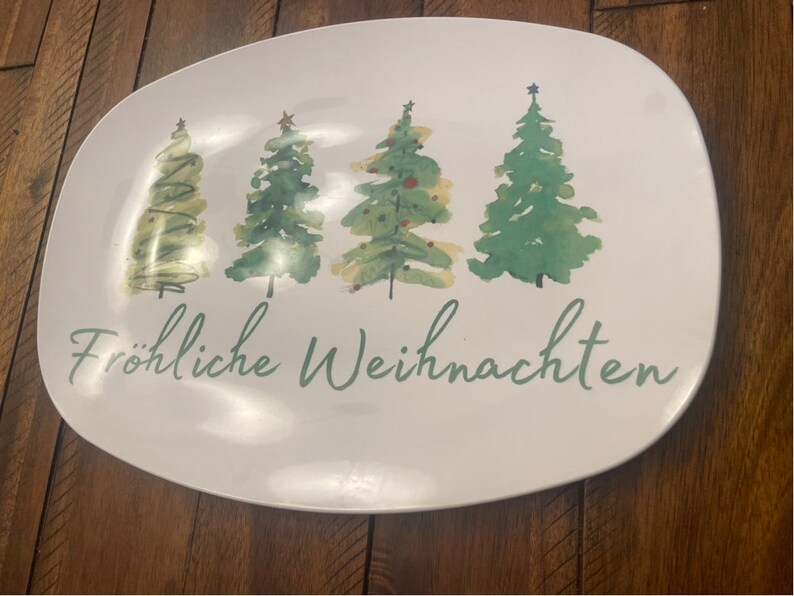 May include: White oval serving platter with a watercolor design of four Christmas trees in varying shades of green. The words "Fr&ouml;hliche Weihnachten" are written in green script below the trees.