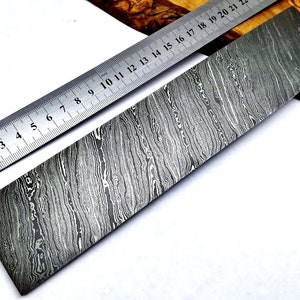 May include: A close-up of a long, rectangular piece of metal with a wavy, patterned surface. The metal is a light grey colour with dark grey lines running through it. A ruler is placed on top of the metal, showing that it is approximately 21 cm long.
