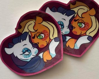 May include: Two heart-shaped stickers featuring a cartoon illustration of a white pony with a purple mane and an orange pony with a blonde mane. The ponies are depicted in a close embrace against a dark purple background with a pink border.