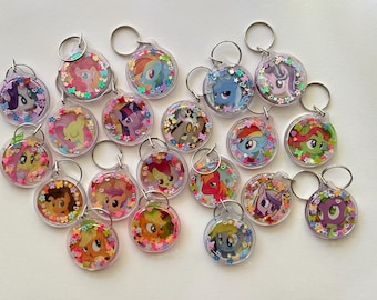 May include: A collection of circular acrylic keychains featuring various cartoon pony characters. Each clear keychain contains colorful, small, shaped glitter pieces including stars, hearts, and flowers surrounding the central character illustration.