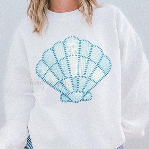 May include: White sweatshirt featuring a light blue patchwork seashell design. The seashell is composed of various blue patterns. The text "FAUX PATCHWORK SEASHELL" is printed in black on the lower right. The model is wearing blue and white striped shorts.
