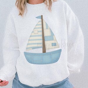 May include: White sweatshirt featuring a blue sailboat design with patchwork details. The sailboat has a brown mast and a blue and white striped sail. The shorts are blue and white striped. The text "FAUX PATCHWORK" is at the bottom.