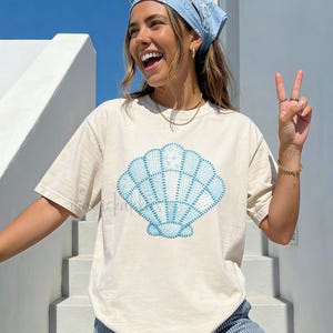 May include: Cream-colored t-shirt featuring a blue seashell design. The graphic is a patchwork style. The text "FAUX PATCHWORK SEASHELL" is printed at the bottom. The person is wearing blue and white striped pants.