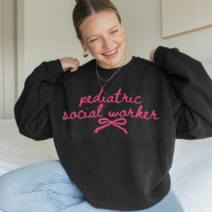 May include: Black sweatshirt with the text "pediatric social worker" in pink script, accented with a pink bow. The sweatshirt is a crew neck style.