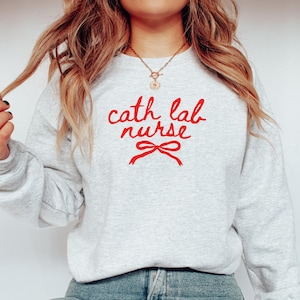 May include: A light gray sweatshirt with the words "cath lab nurse" in red script, and a red bow graphic. The sweatshirt is made of a soft material and has long sleeves.