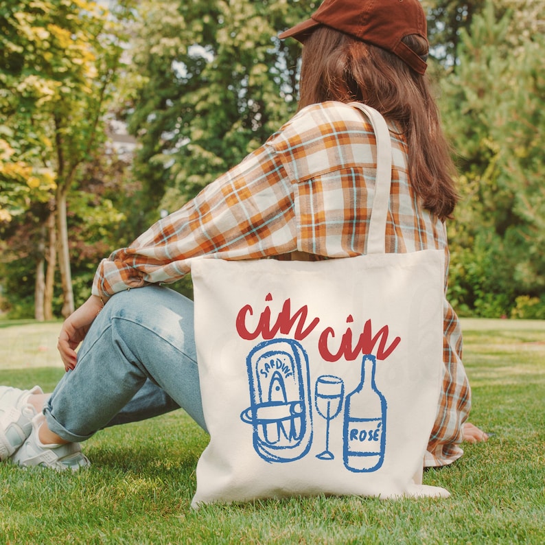 A natural colored cotton canvas tote bag. Design large in center of bag. Design says cin cin in fancy print in deep bright red all lowercase and curved on top of elements below. Elements pictured are thick sketched doodles of tinned sardines and a rose bottle with a wine glass in the center of both elements. Elements color is in bright coastal blue. Elements have a slight distressed look to them. For the fisherman aesthetic, Italian and mediterranean summer aesthetic, and sardine lovers.