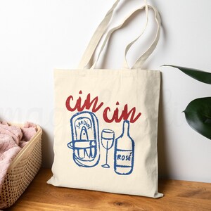 A natural colored cotton canvas tote bag. Design large in center of bag. Design says cin cin in fancy print in deep bright red all lowercase and curved on top of elements below. Elements pictured are thick sketched doodles of tinned sardines and a rose bottle with a wine glass in the center of both elements. Elements color is in bright coastal blue. Elements have a slight distressed look to them. For the fisherman aesthetic, Italian and mediterranean summer aesthetic, and sardine lovers.
