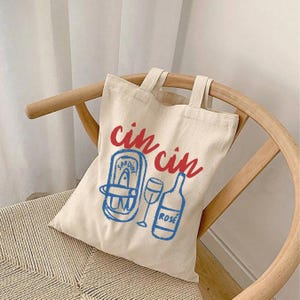 A natural colored cotton canvas tote bag. Design large in center of bag. Design says cin cin in fancy print in deep bright red all lowercase and curved on top of elements below. Elements pictured are thick sketched doodles of tinned sardines and a rose bottle with a wine glass in the center of both elements. Elements color is in bright coastal blue. Elements have a slight distressed look to them. For the fisherman aesthetic, Italian and mediterranean summer aesthetic, and sardine lovers.