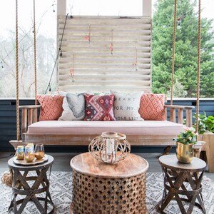 May include: A porch swing with a pink cushion and colorful pillows. The swing is hanging from the ceiling with rope. There are two small tables with intricate designs and a larger table with a woven lantern on top. The floor is covered in a white and gray patterned rug.