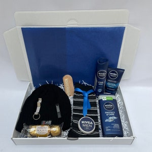 May include: A white gift box filled with a blue and black beanie, a pair of striped socks, a wooden comb, a small jar of cream, a bottle of body wash, a bottle of deodorant, and a package of chocolates. The box is lined with white shredded paper.