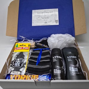May include: A white gift box filled with various items, including a black and white striped sock, a black car air freshener, a wooden brush, a black and white chocolate bar, and two black bottles of Lynx body spray.
