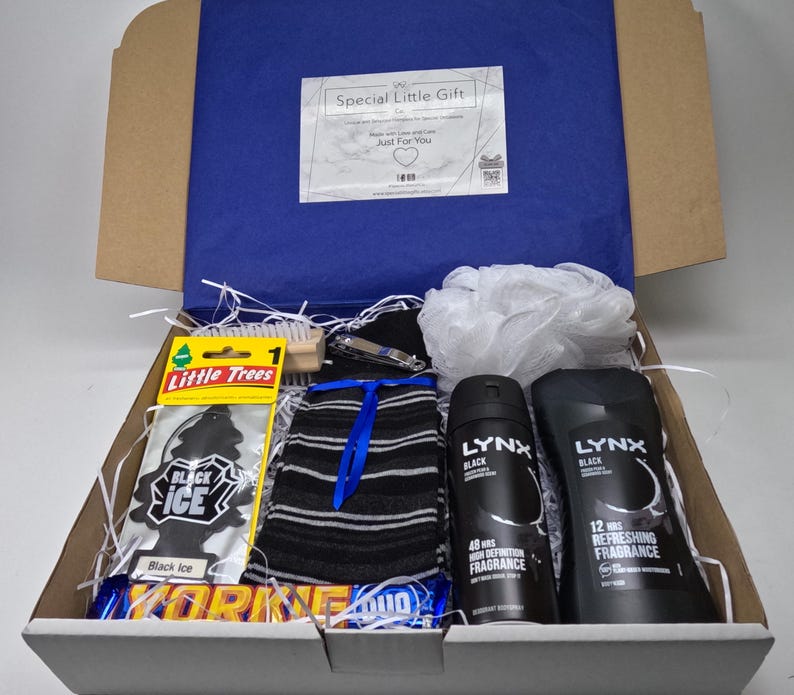 May include: A white gift box filled with various items, including a black and white striped sock, a black car air freshener, a wooden brush, a black and white chocolate bar, and two black bottles of Lynx body spray.