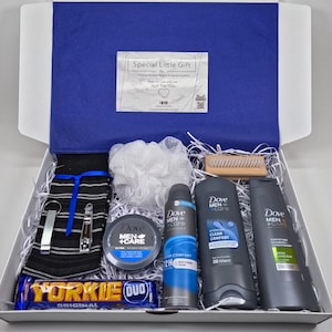 May include: A white gift box filled with a variety of grooming products, including a white washcloth, a black and white striped towel, a wooden brush, a chocolate bar, and three Dove Men+Care products: a body wash, a deodorant, and a shaving cream. The box is lined with white shredded paper.