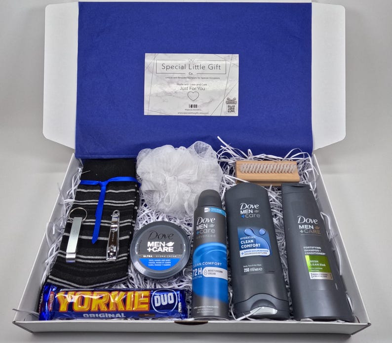 May include: A white gift box filled with a variety of grooming products, including a white washcloth, a black and white striped towel, a wooden brush, a chocolate bar, and three Dove Men+Care products: a body wash, a deodorant, and a shaving cream. The box is lined with white shredded paper.